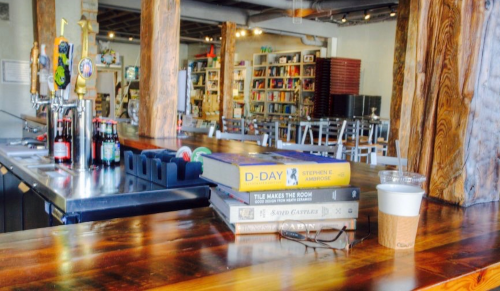 A cozy café interior with a wooden bar, stacked books, and a coffee cup, surrounded by shelves of books.