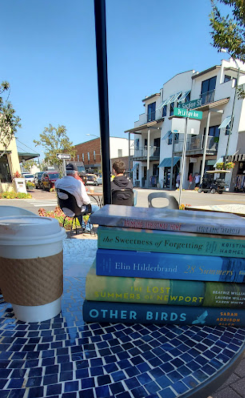 A coffee cup sits on a mosaic table with stacked books, while people relax in a sunny outdoor setting.