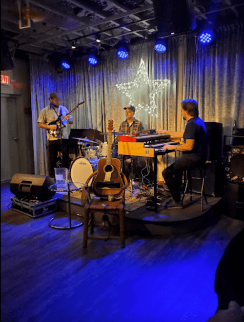 A band performs on stage with guitar, drums, and keyboard under blue lights and a star decoration.