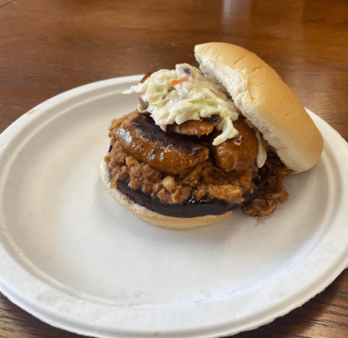 A sandwich with pulled pork, sausage, and coleslaw on a bun, served on a paper plate.