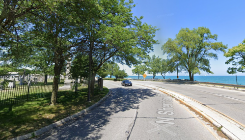 A scenic view of a curved road lined with trees, leading to a lake with a clear sky in the background.