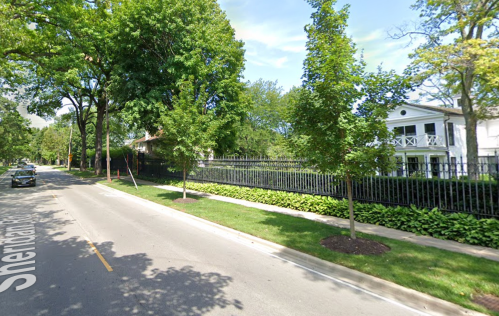 A tree-lined street with a black fence and a white house in the background, surrounded by greenery.