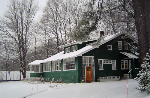 A green house with a snow-covered yard, surrounded by trees in winter. Snow blankets the ground and rooftops.