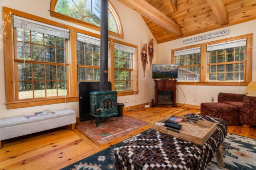 Cozy living room with wooden beams, a wood stove, large windows, and rustic decor, overlooking a forested area.