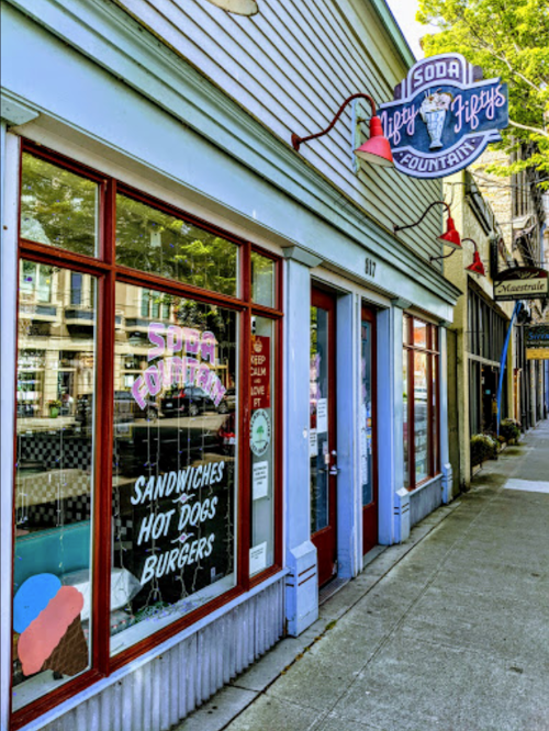 Exterior of a retro soda fountain shop with colorful signage, featuring windows displaying menu items like sandwiches and burgers.