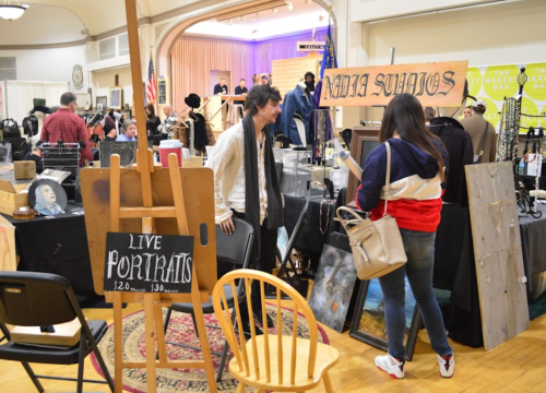 A bustling art fair with vendors, live portrait sign, and visitors browsing various artworks and crafts.