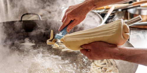 A chef's hands squeeze dough from a pastry bag into bubbling oil, creating fresh fried pastries. Steam rises around them.