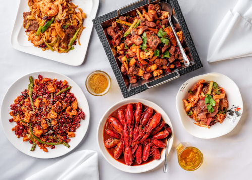 A spread of various Asian dishes, including stir-fried noodles, spicy meats, and seafood, with drinks on a white tablecloth.