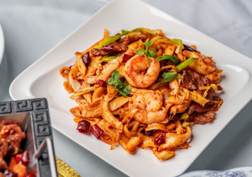 A plate of stir-fried noodles with shrimp, vegetables, and spices, garnished with green onions and cilantro.