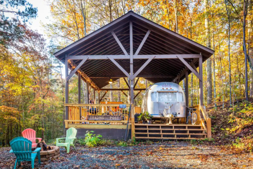 A cozy Airstream trailer under a wooden pavilion, surrounded by autumn trees and outdoor seating.
