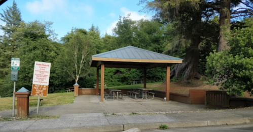 A covered picnic area with tables in a park, surrounded by trees and grass, with a sign nearby.