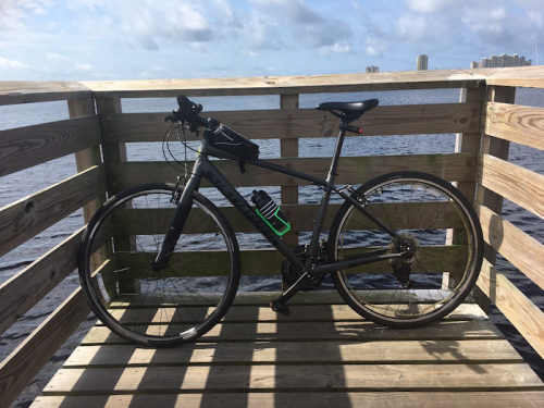A black bicycle rests on a wooden dock overlooking a calm body of water under a partly cloudy sky.