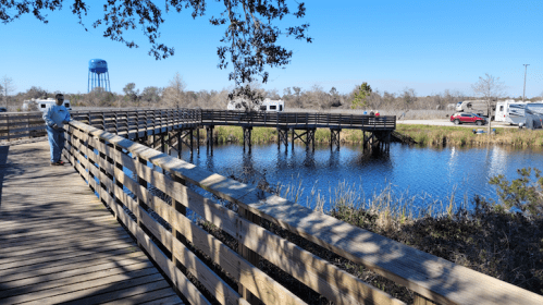 A wooden boardwalk curves over a calm waterway, with a water tower and RVs visible in the background under a clear blue sky.