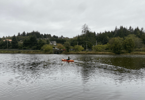 A person paddles a kayak on a calm river surrounded by trees and houses under a cloudy sky.