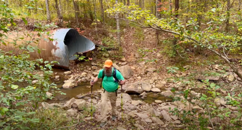 A hiker in a green shirt and orange hat navigates rocky terrain near a creek and a metal pipe in a wooded area.