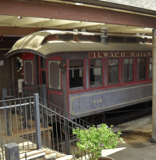 Historic Ilwaco Railway train car displayed under a covered structure, with a small plant nearby.