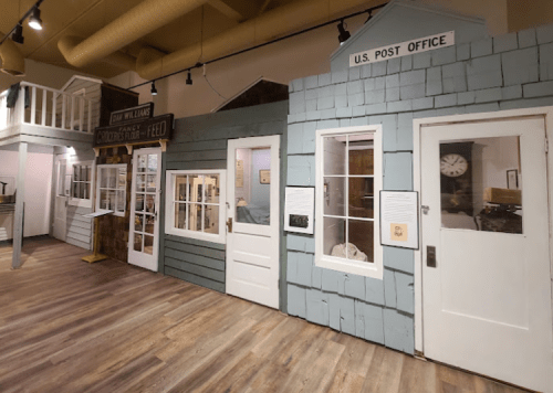 A display of vintage storefronts, including a post office, with wooden facades and large windows in a museum setting.