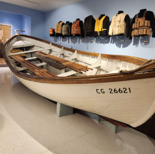 A vintage white boat displayed indoors, with life vests hanging on the wall behind it.