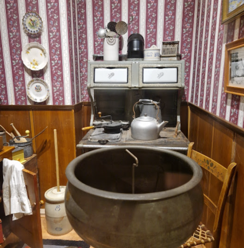 A vintage kitchen with a stove, kettle, and utensils, featuring patterned wallpaper and rustic decor.