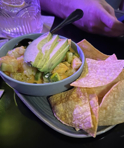 A bowl of creamy dip topped with avocado slices, served with crispy tortilla chips on a dark table.