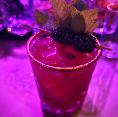 A close-up of a cocktail garnished with mint and blackberries, set against a purple-lit background.