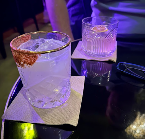 Two cocktails on a table, one with a rim of red salt and ice, the other clear with a lemon slice, on coasters.