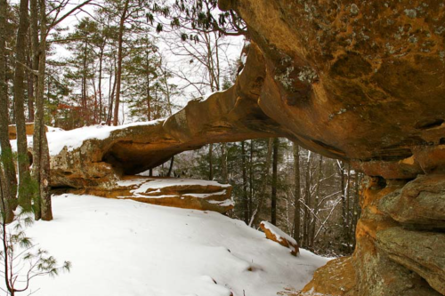 A snow-covered rock formation arches over a forested area, surrounded by trees in winter.