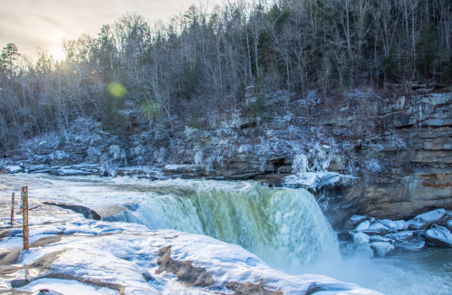 A frozen waterfall cascades over rocky cliffs, surrounded by trees and icy terrain under a bright winter sky.