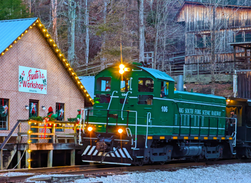 A green locomotive passes by a festive workshop decorated for the holidays, surrounded by trees and rustic buildings.