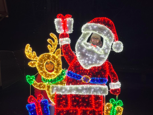 Two people pose inside a brightly lit Christmas display featuring Santa and a reindeer, surrounded by colorful decorations.