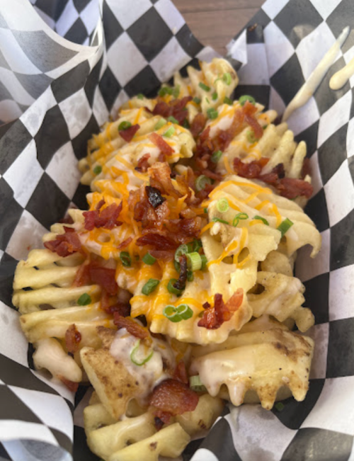 A serving of waffle fries topped with cheese, bacon bits, and green onions in a checkered paper basket.