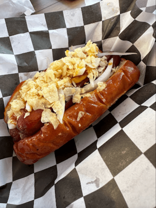 A hot dog in a bun topped with onions and crushed potato chips, served in a black and white checkered paper.