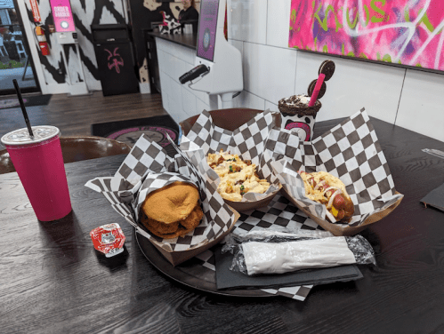 A table with three food items in checkered baskets, a pink drink, and a dessert, set in a colorful diner.