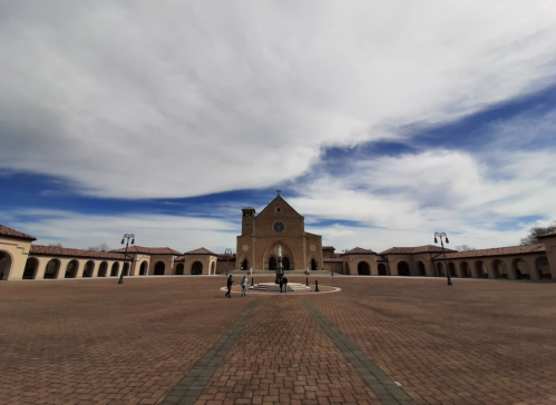 A spacious plaza with a large building at the center, surrounded by arches and a blue sky with clouds.