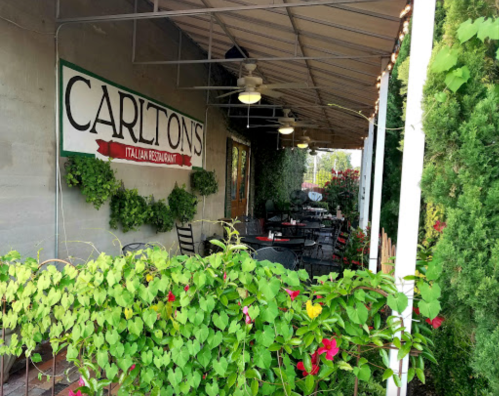 Exterior of Carlton's Italian Restaurant, featuring outdoor seating surrounded by greenery and a welcoming sign.