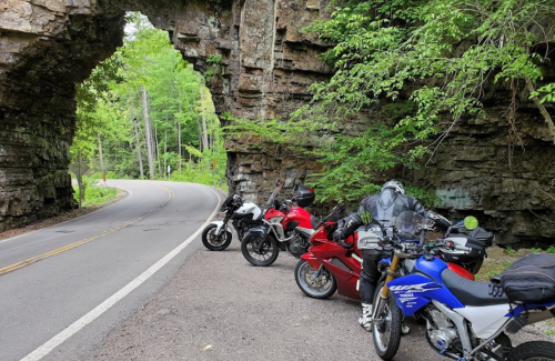 Four motorcycles parked under a rocky archway, surrounded by lush greenery along a winding road.