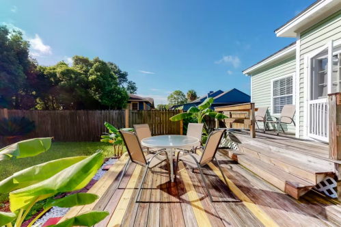 A sunny backyard patio with a round table and chairs, surrounded by greenery and a wooden deck.