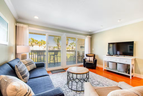 Cozy living room with a blue sofa, coffee table, TV, and large windows overlooking a patio with palm trees.