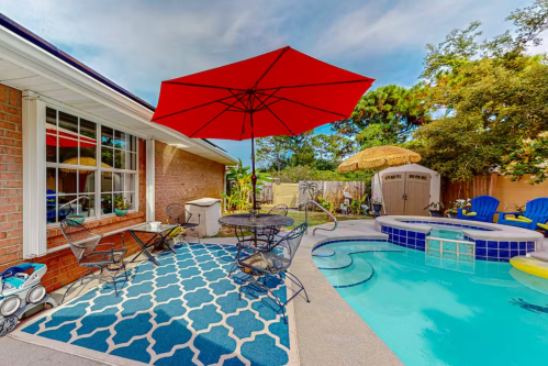 A backyard pool area featuring a blue rug, red umbrella, seating, and a hot tub surrounded by greenery.