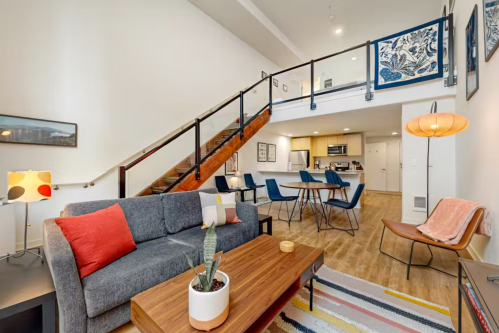 Modern living room with a gray sofa, wooden coffee table, and a staircase leading to a dining area and kitchen.