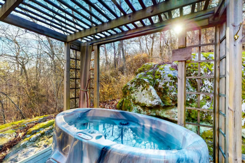 A hot tub on a wooden deck surrounded by trees and rocks, with sunlight filtering through a pergola.