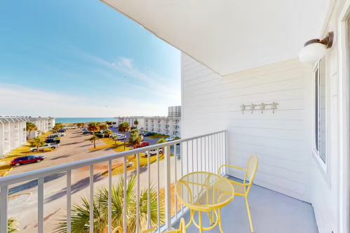 A balcony with yellow furniture overlooks a parking lot and the ocean in the distance under a clear blue sky.