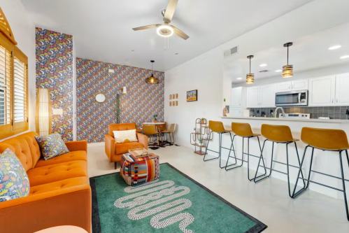 Bright living room with orange sofa, patterned wallpaper, and a kitchen bar with stools. Cozy and modern decor.