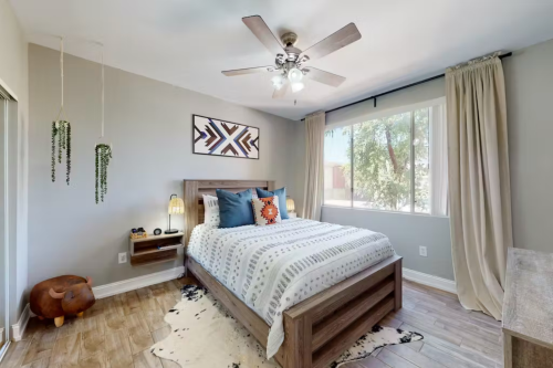 Cozy bedroom with a wooden bed, patterned bedding, large window, and decorative plants. Natural light fills the space.