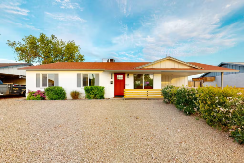 A single-story house with a red door, surrounded by gravel and greenery, under a blue sky with scattered clouds.