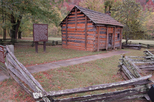 A rustic log cabin surrounded by a wooden fence, with informational signs nearby and trees in the background.