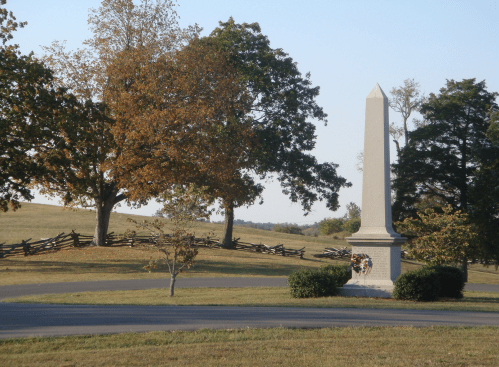 A tall stone monument surrounded by trees and a grassy field, with a wooden fence in the background.