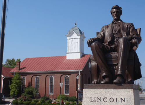 A bronze statue of Abraham Lincoln sits in front of a historic brick building with a white clock tower.