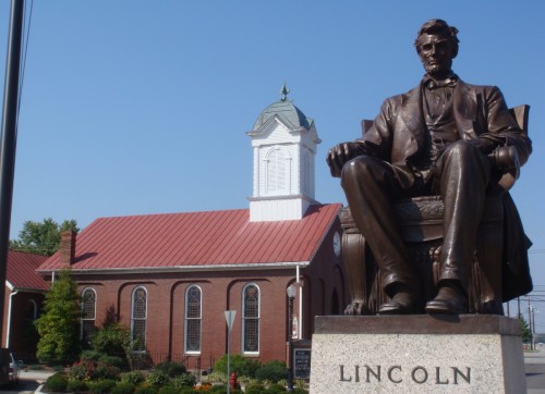 Statue of Abraham Lincoln seated in front of a historic brick church with a white steeple against a clear blue sky.
