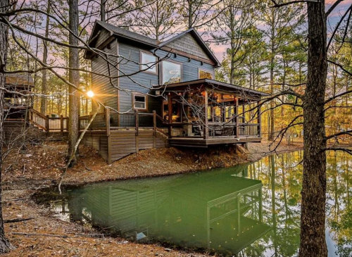 A cozy cabin by a serene pond, surrounded by trees and bathed in warm sunset light.